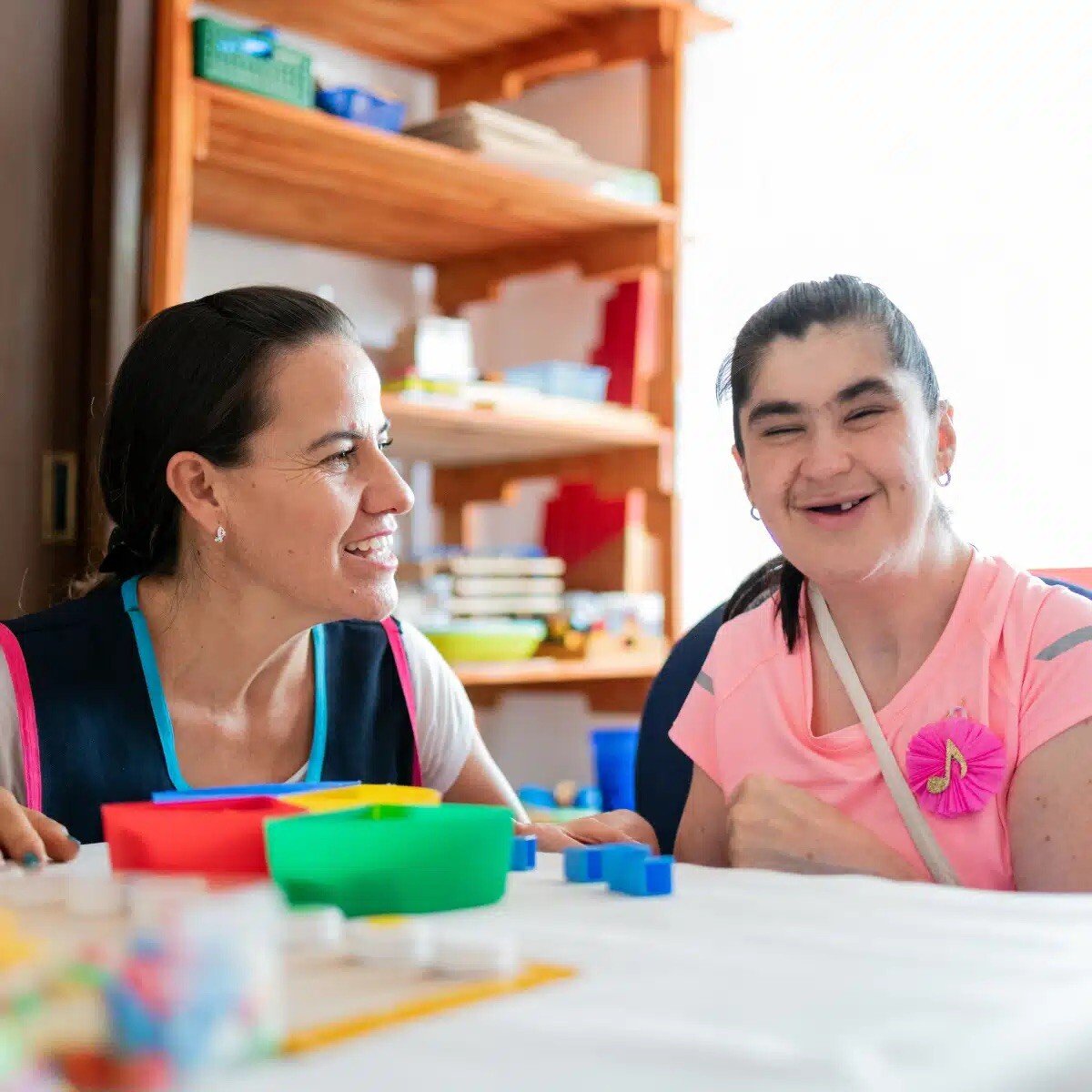 A woman and a young girl sit at a table, smiling, playing with building blocks.